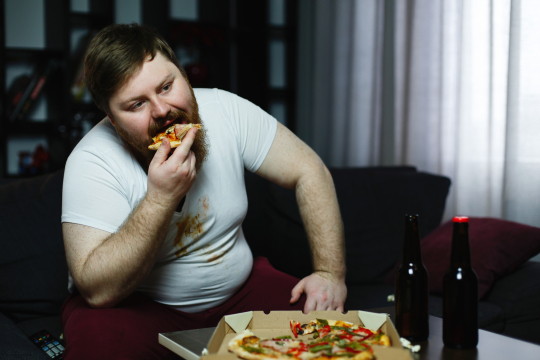 Ugly fat man eats pizza sitting on the sofa
